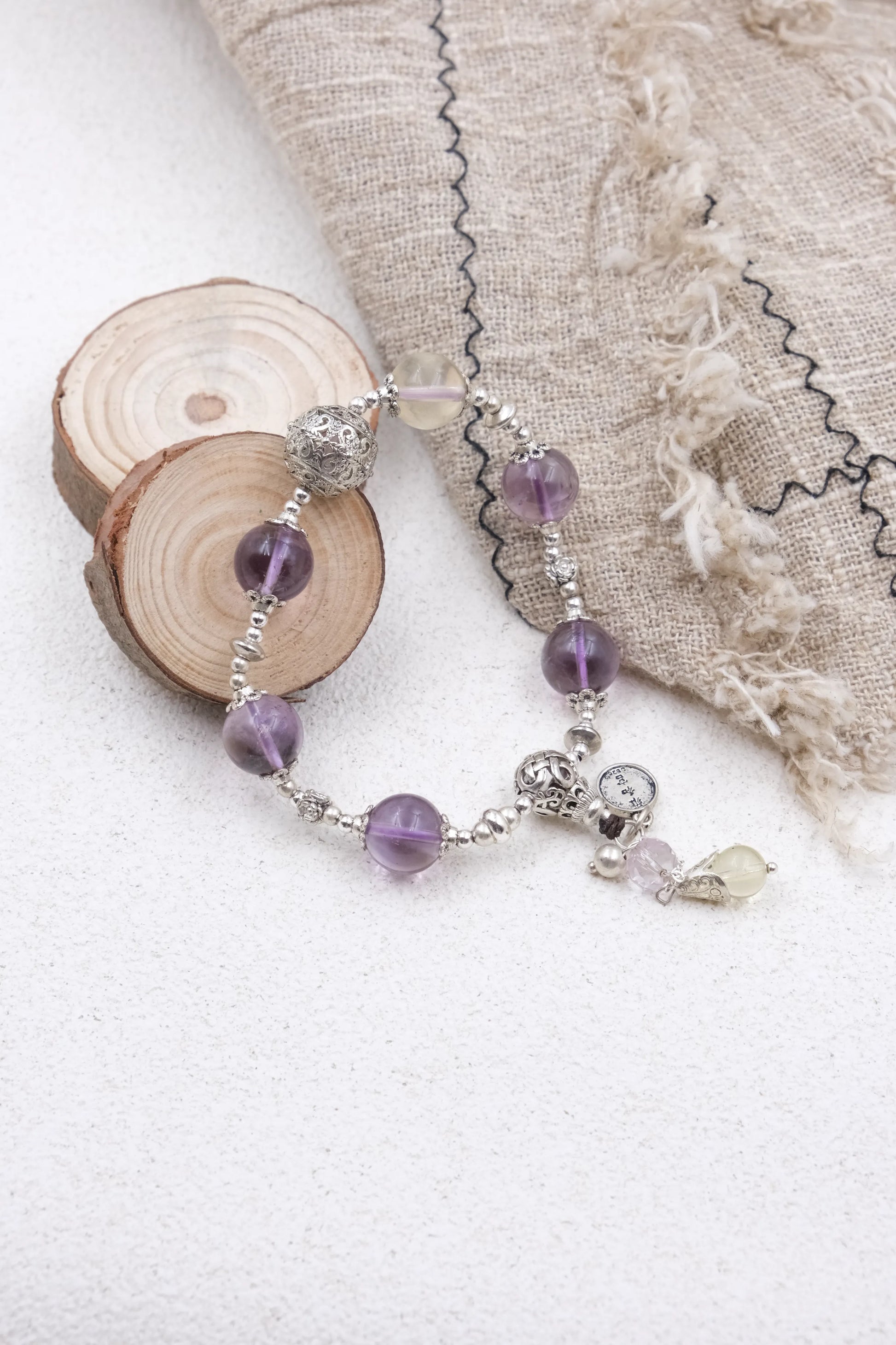 Vajra Bell Crystal Energy Bracelet Beaded bracelet with purple and clear beads on a white surface with fabric and wood slice in the background