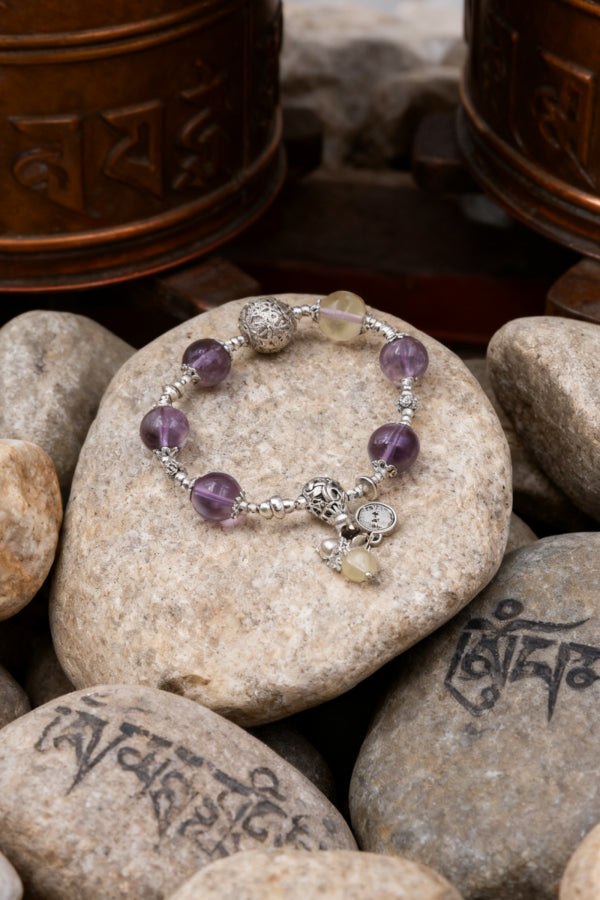 A bracelet with amethyst beads and silver accents, featuring charms, placed on a rock, with prayer wheels and inscribed stones in the background.