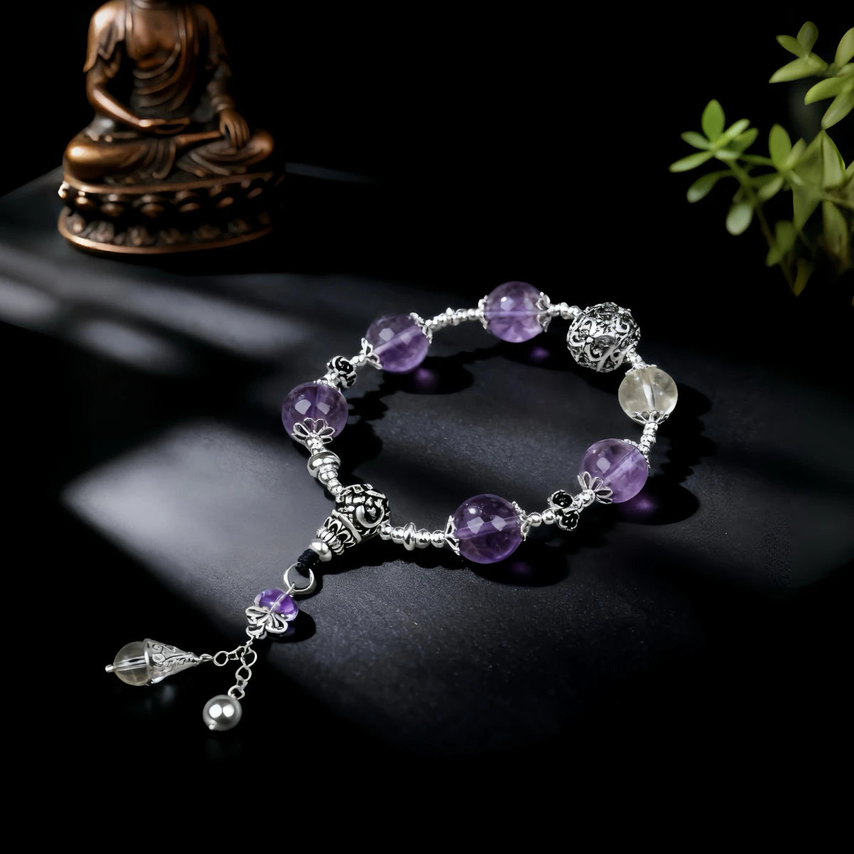 Purple beaded bracelet with silver charms on a dark surface, with a small statue and plant in the background.