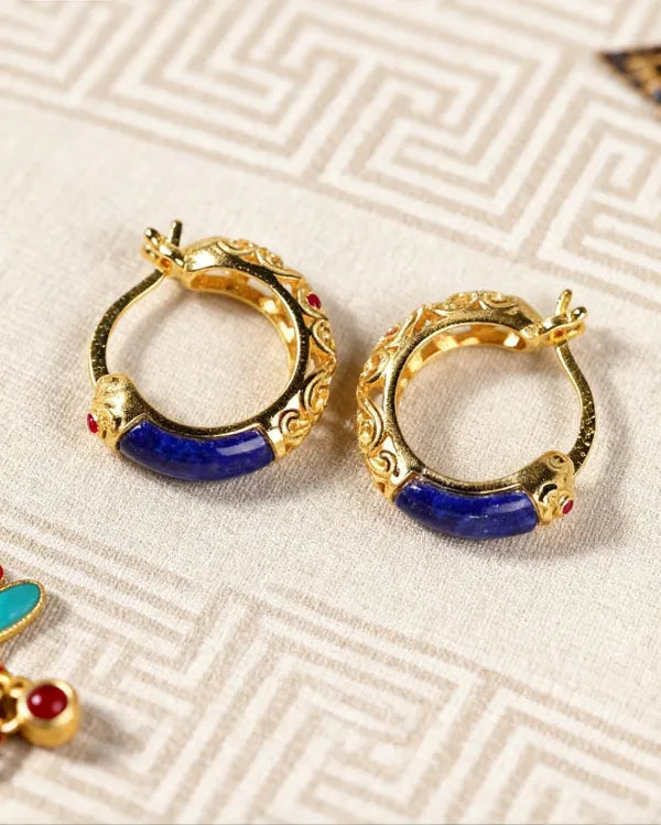 Vintage gold hoop earrings with lapis lazuli and red gem accents, featuring intricate carvings and spiritual oriental design on patterned fabric background.