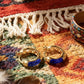 Golden hoop earrings with blue lapis lazuli inlays on a colorful patterned rug with fringes, beside a feather and a beaded bracelet.