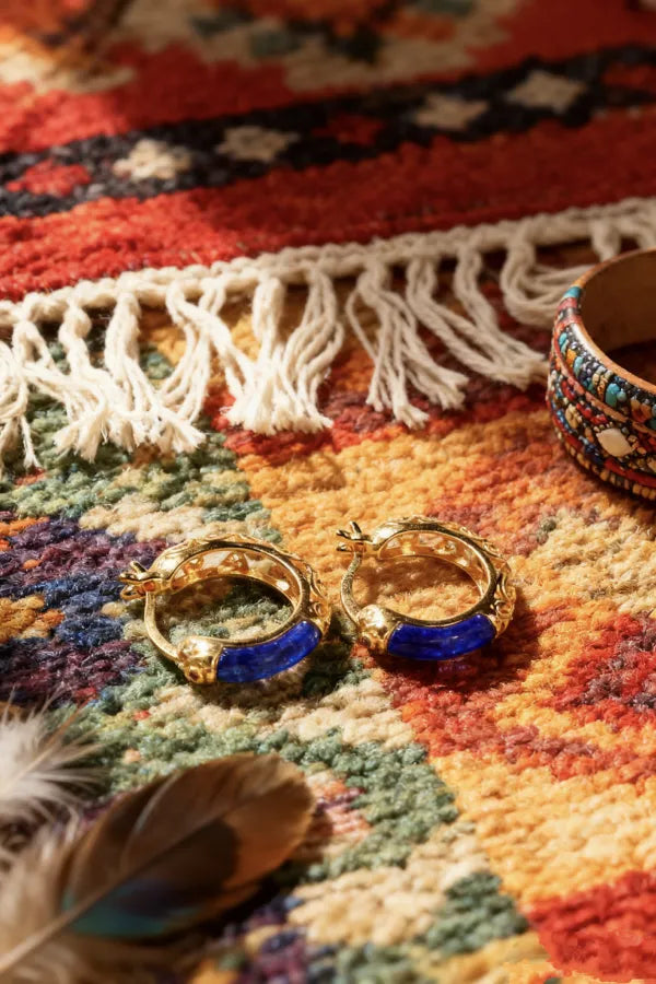 Golden hoop earrings with blue lapis lazuli inlays on a colorful patterned rug with fringes, beside a feather and a beaded bracelet.