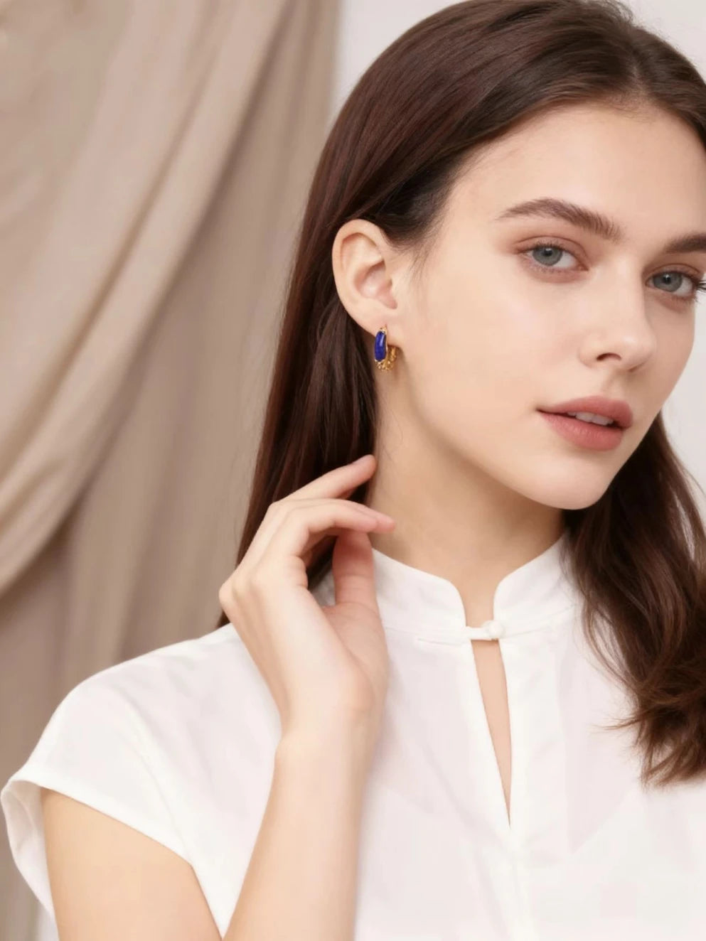 A woman wearing elegant golden hoop earrings with blue lapis lazuli accents, dressed in a white top, against a soft neutral-toned curtain background.