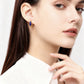 A woman wearing elegant golden hoop earrings with blue lapis lazuli accents, dressed in a white top, against a soft neutral-toned curtain background.