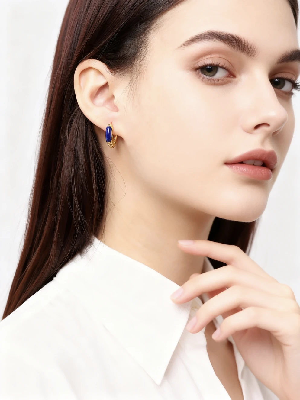 A woman wearing elegant golden hoop earrings with blue lapis lazuli accents, dressed in a white top, against a soft neutral-toned curtain background.