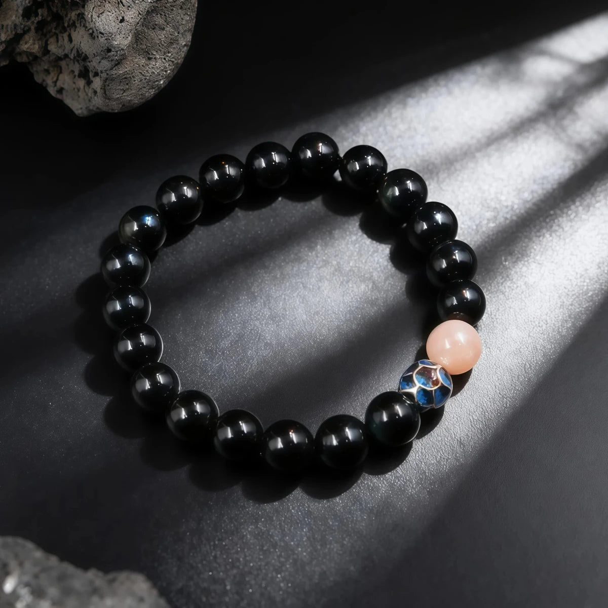 The black bracelet displayed under striped lighting shadows, highlighting the pink stone and blue bead.