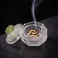 A clear carved glass incense burner with a matching lid and green lotus-shaped knob, shown open with a smoking incense stick inside on a dark textured surface.