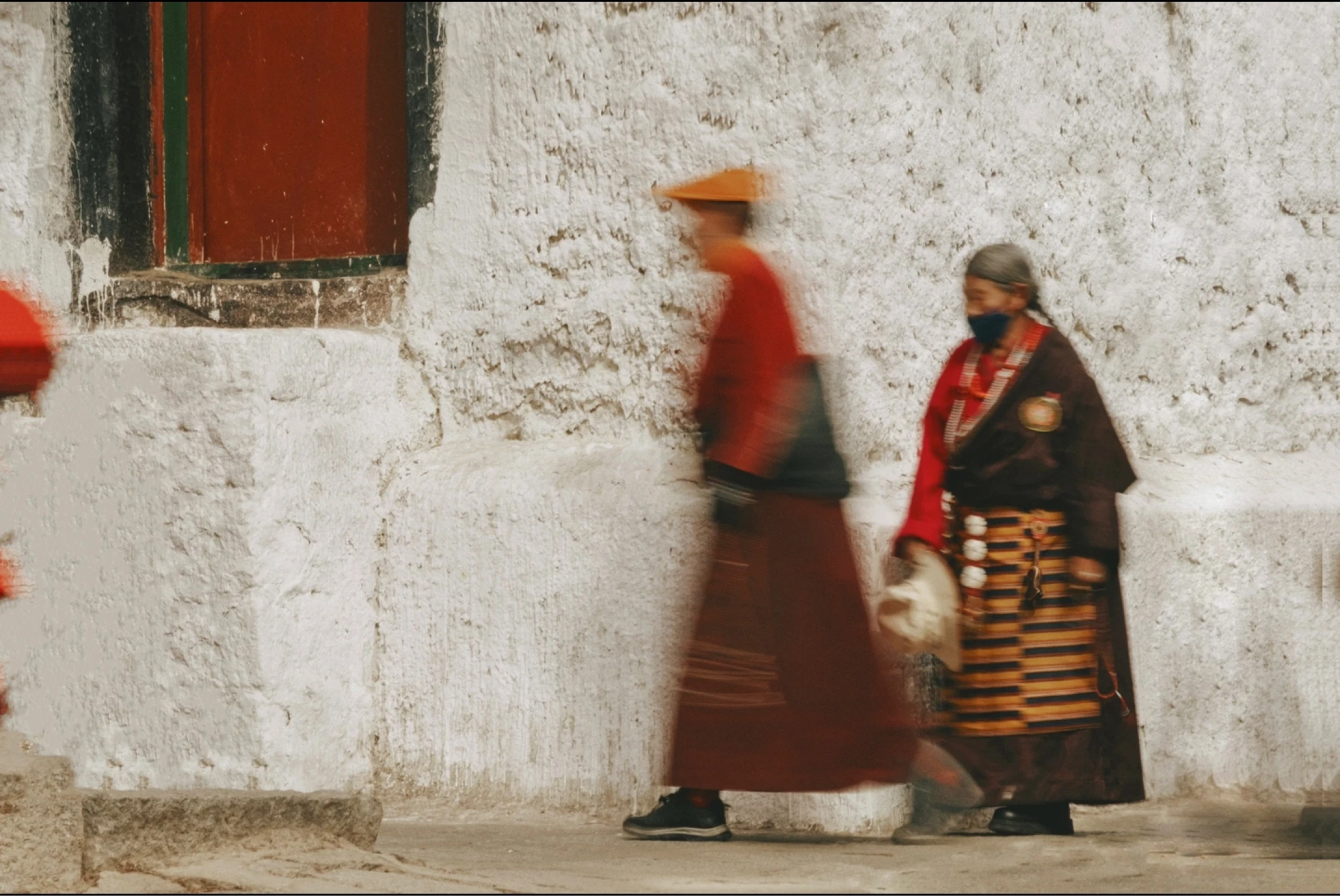Two tibetan people in traditional attire walking past a white wall with a red door.